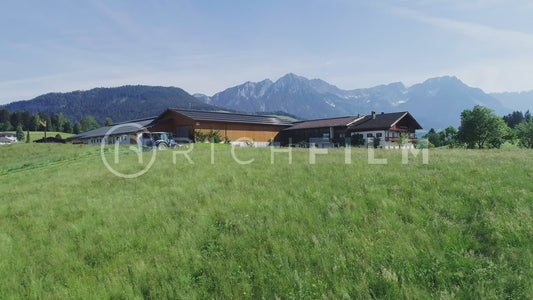 Aerial view of a tractor in the field mowing next to a farm with the Alps in the backgroun