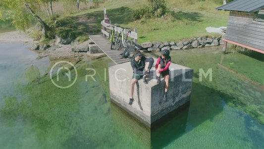 Aerial view of two cyclists sitting next to a hut on a jetty by a lake on a sunny day