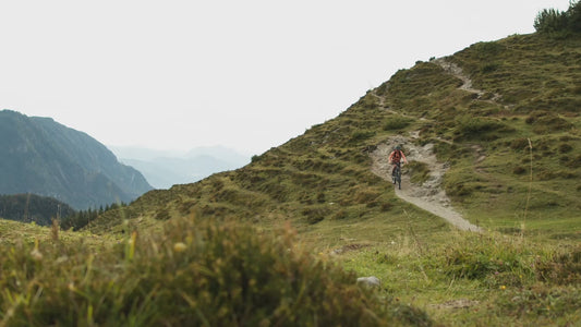 Slow motion shot of a mountain biker riding over a hill with a view of the mountains and clouds