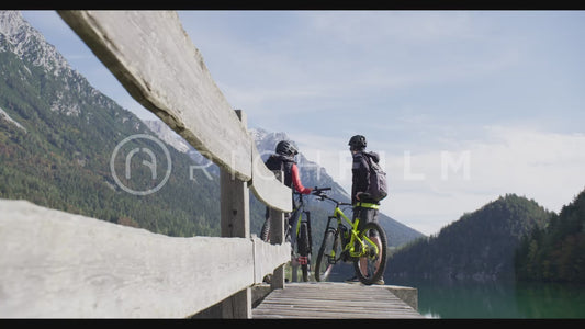 Shot of bikers high-fiving on a pedestrian bridge with the Alps and a lake in the background