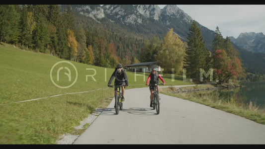 shot of mountain bikers riding on a road next to a lake with mountains in the background