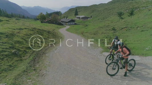 Aerial shots of mountain bikers riding a gravel road through mountain houses with the alps in the background